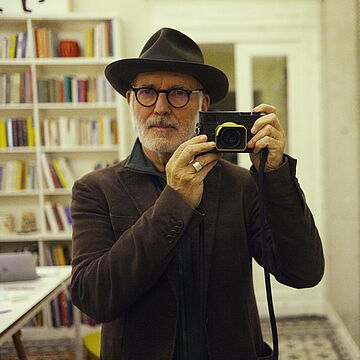 Ludovico mit Hut und einem Fotoapparat in den Händen steht in einem Zimmer mit einer Bücherwand. 